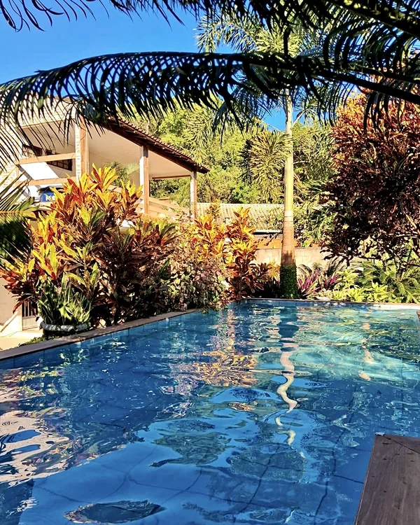 Reflexos na água da piscina, com jardim tropical e varanda ao fundo.