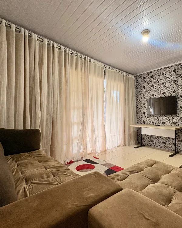 Sala com sofá, tapete, cortinas claras e televisão na parede.