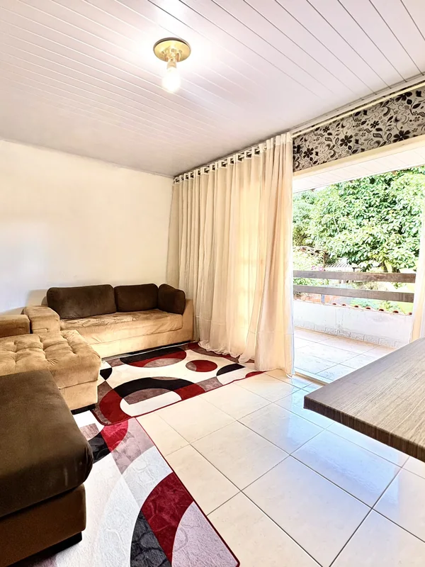 Sala com sofás, cortinas claras e abertura para a varanda com vista para o verde.