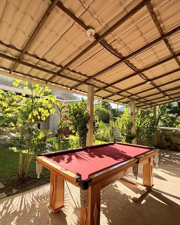 Mesa de sinuca com tecido vermelho em uma área coberta junto ao jardim.